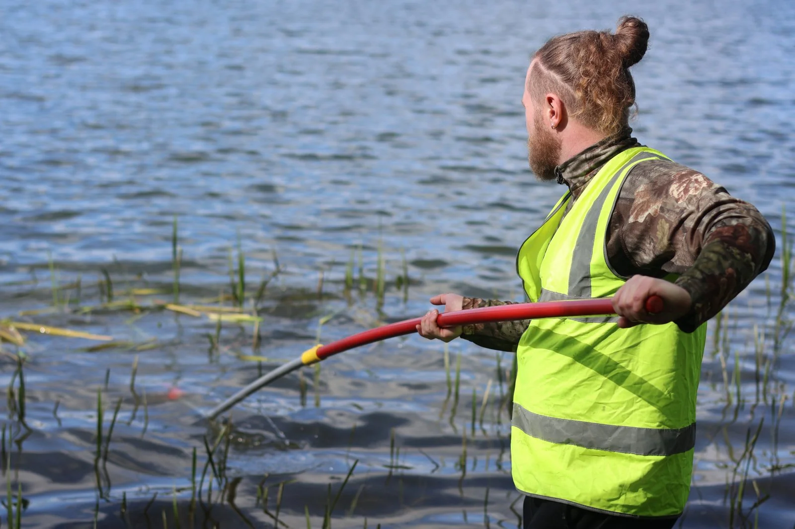 A volunteer in a high-vis vest using a long-handled tool to clear weed from a lake margin. The kind of physical work anglers do at clubs across the country, almost none of it captured in any formal sector statistic.