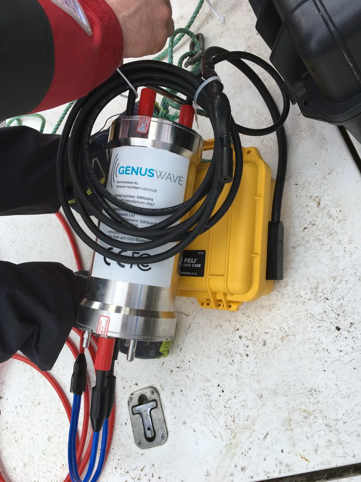 Photograph of an actual Genuswave A1 unit on the deck of a research vessel, alongside its yellow Peli 1200 transit case. The stainless steel cylindrical housing is visible with the Genuswave label, cabling coiled around it, ready for deployment. Real working equipment in deployment context, not a schematic.