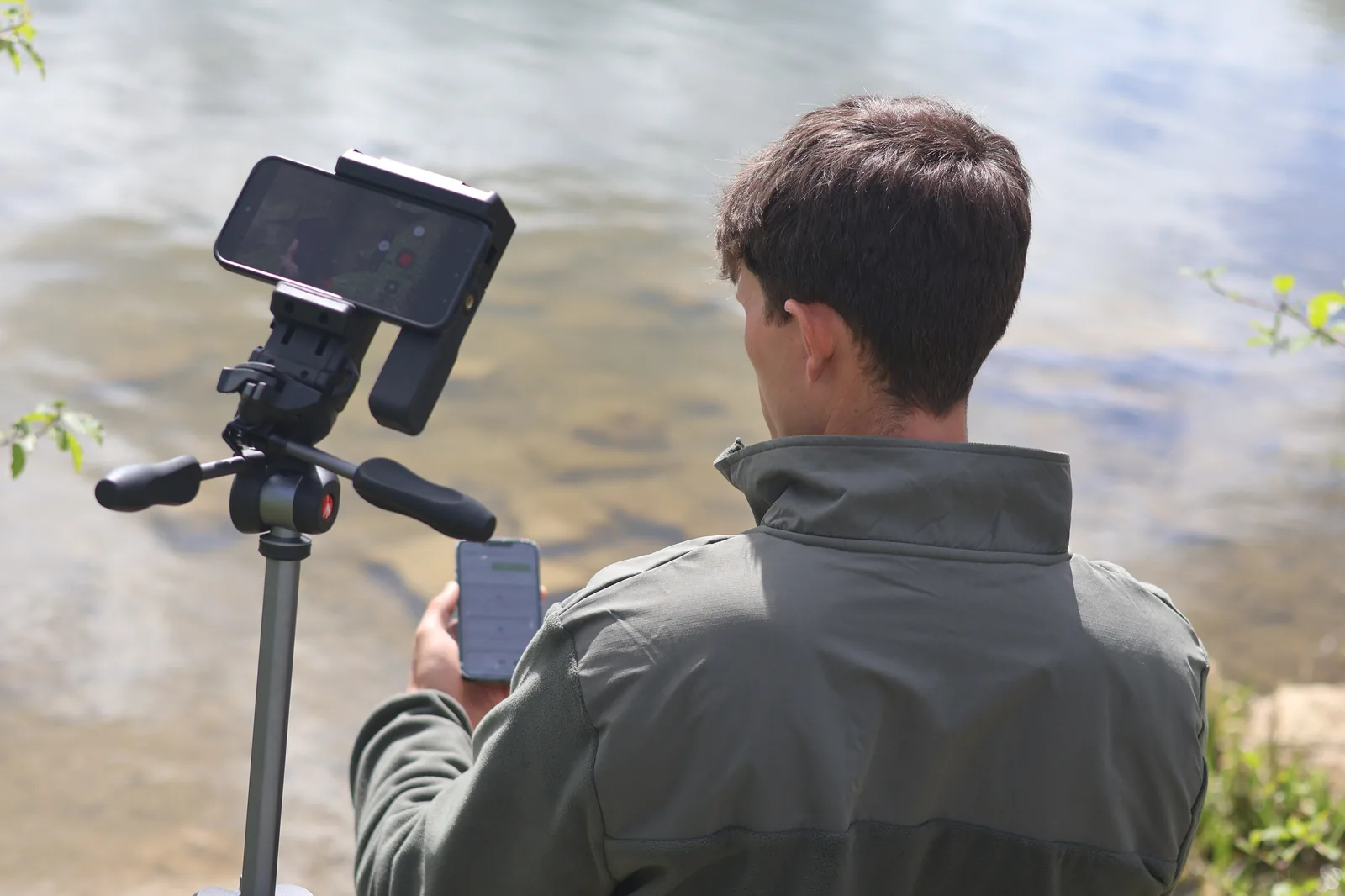 A volunteer at a UK lakeside recording biodiversity through Hydroscape — phone on a tripod framing the water, a second phone in hand showing the submission form open